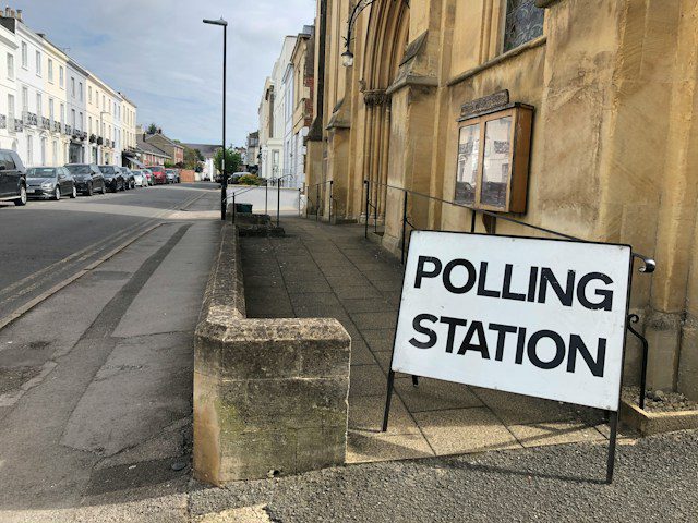 Sign on the Pavement shows 'Polling Station' against background of town centre.